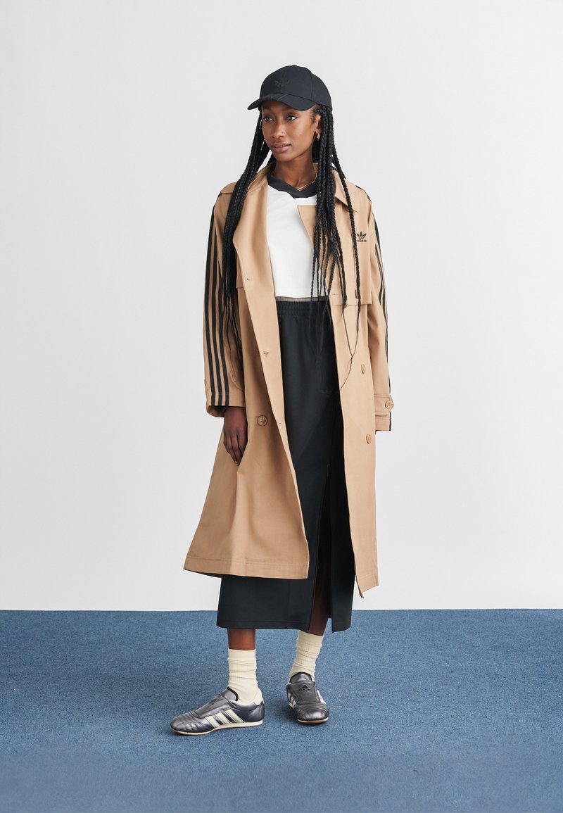Beige trenchcoat med sorte striber, hvid top, sort nederdel og grå sneakers med hvide detaljer. En sort kasket og sokker fuldender looket.