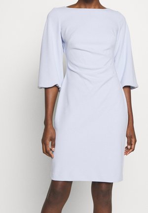 Femme portant une robe bleu clair longueur genou avec des manches ballon trois-quarts et un col bateau, debout devant un fond uni.