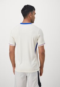 Maglia sportiva bianca con colletto blu e accenti blu, maniche corte, tessuto texturizzato, con strisce rosse e blu ai polsini.