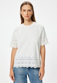 Camiseta blanca de manga corta con detalles de encaje en las mangas y el dobladillo, con un cuello redondo y un ajuste relajado.