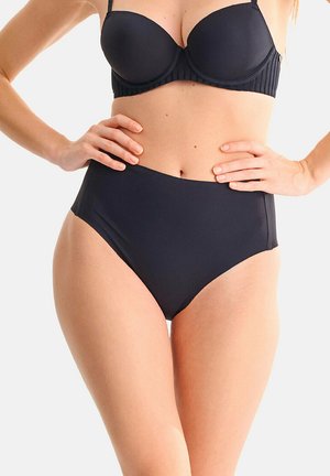 Femme portant un bas de bikini noir taille haute et un haut de bikini noir avec armatures, les mains posées sur les hanches sur un fond uni.