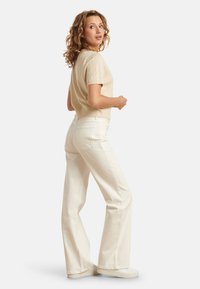 Beige gebreid T-shirt met korte mouwen en een textuurpatroon, hoge taille witte flared jeans en witte sneakers, weergegeven vanuit een iets gedraaide hoek.