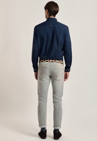 Camisa de botones azul marino de manga larga, combinada con jeans gris claro y un cinturón estampado. Zapatos marrones y puños remangados completan el look.