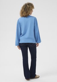 Svetlo modra dolgi rokav s nabranimi rokavi, v kombinaciji z temno modrimi širokimi kavbojkami in svetlimi čevlji. Preprost, sproščen dizajn s udobnim krojem.