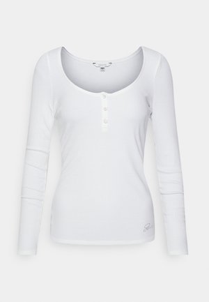 White long-sleeve top with a scoop neckline, ribbed texture, and three decorative buttons at the front. Subtle logo detail on the bottom.