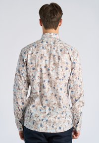 Mees näoga eemale, kandes beeži värvi lillelist pika varrukaga särki siniste ja pruuni lillemustritega, tumedate pükstega, ühtlase taustaga.