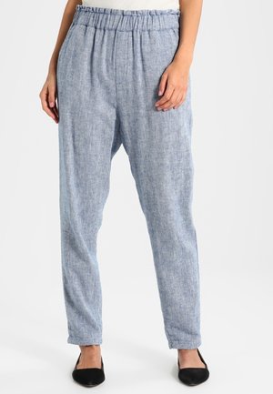 Femme portant un pantalon en lin bleu clair taille haute avec une ceinture élastique et des jambes fuselées, associé à des ballerines noires pointues.