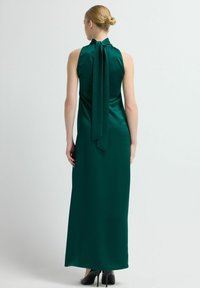 Dlouhé, bezrukávové šaty z tmavě zeleného saténu; mají vysoký límec s mašlí a sahají až k zemi, ukazují hladkou texturu.