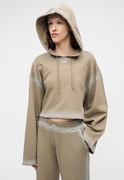 Γυναίκα με μπεζ κοντό φούτερ με κουκούλα και ταιριαστό παντελόνι φόρμας, και τα δύο με το κείμενο "OFF", στέκεται μπροστά σε απλό φόντο.