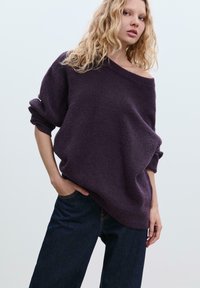 Übergroßer lila Pullover mit schulterfreiem Design, gerippten Bündchen und weicher Textur, kombiniert mit dunkelblauen Jeans.