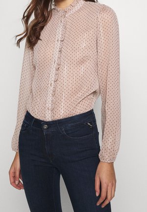 Vrouw draagt een doorschijnende lichtroze blouse met lange mouwen, zwarte polkadots en een gerimpelde knoopsluiting, gecombineerd met donkerblauwe aansluitende jeans.