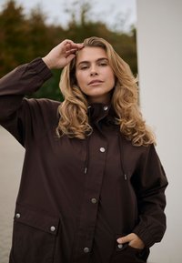 Femme aux longs cheveux blonds bouclés portant une veste marron foncé, avec une main touchant sa tête et l'autre dans sa poche, à l'extérieur.