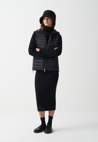 Zwarte gewatteerde vest met een capuchon over een zwart ribbel midi-jurk. Zwarte bucket hat en schoenen maken de look compleet. Gevarieerde texturen overal.