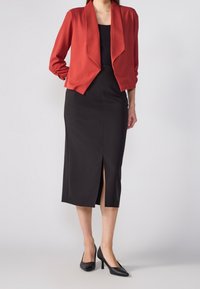 Chaqueta roja abierta por delante sobre un vestido negro ajustado con una abertura alta en el muslo. El conjunto tiene una textura suave y una silueta clásica, combinado con tacones negros.