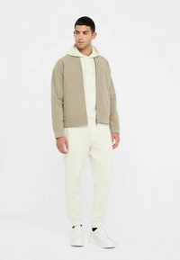 Beige Bomberjacke mit Reißverschluss vorne, zwei seitlichen Taschen und gerippten Bündchen, darüber getragen ein hellfarbener Hoodie, kombiniert mit weißen Jogginghosen.