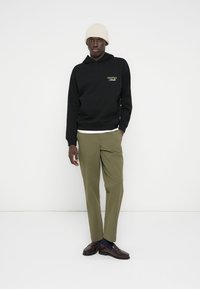 Μαύρο φούτερ με κουκούλα και μπροστινή τσέπη, με χρυσά γράμματα «ORIGINALS» στο στήθος. Ελαιδί στενά παντελόνια, καφέ παπούτσια και μπεζ πλεκτός σκούφος.