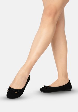 Benen met zwarte ballerina's met kleine strikjes, gekruist staand op een effen lichte achtergrond.