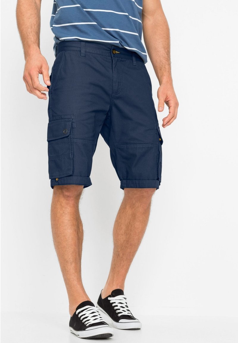 Pantaloni cargo blu realizzati in tessuto resistente, dotati di molteplici tasche, risvolti e una vestibilità dritta. Abbinati a scarpe da ginnastica in tela nere.