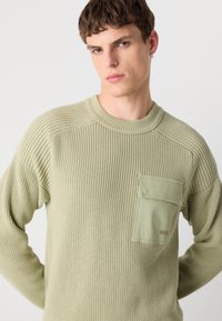 Pull en maille côtelée vert clair avec un col rond, comportant une poche texturée et un logo de marque discret sur le devant.