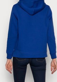 Felpa con cappuccio blu dal tessuto morbido, polsini a costine e tasca a marsupio; abbinata a jeans scuri e aderenti.