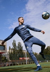 Atlet v temno modrem Puma trening kompletu s turkiznimi detajli, ki brca nogometno žogo na igrišču. V ozadju so stavbe in drevesa.