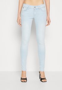 Jean skinny bleu clair avec cinq poches, fermeture à bouton à l'avant, texture lisse et détails de couture minimalistes, associés à des sandales à talons noires.