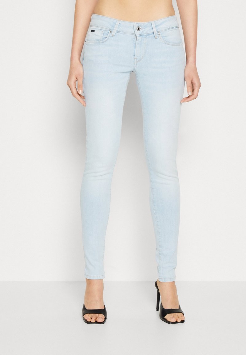 Jean skinny bleu clair avec cinq poches, fermeture à bouton à l'avant, texture lisse et détails de couture minimalistes, associés à des sandales à talons noires.