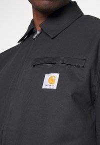 Černá pracovní bunda z odolné látky, vybavená zipovou kapsou na hrudi a výrazným logem Carhartt v žluté barvě na bílém pozadí.