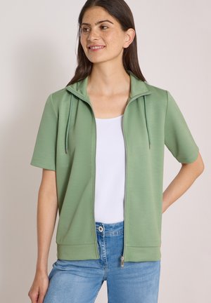 Femme portant une veste zippée à manches courtes vert clair sur un haut blanc et un jean bleu, debout avec une main sur la hanche, regardant sur le côté et souriant.