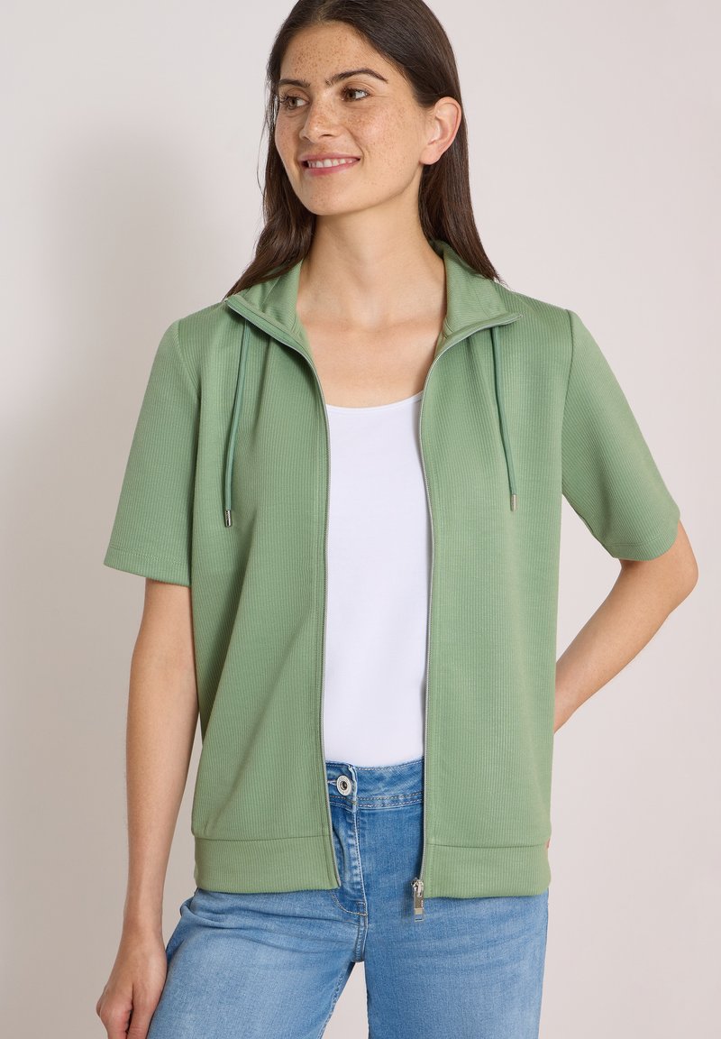 Femme portant une veste zippée à manches courtes vert clair sur un haut blanc et un jean bleu, debout avec une main sur la hanche, regardant sur le côté et souriant.