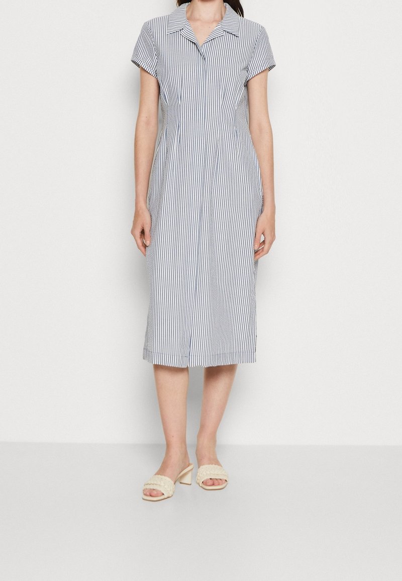 Femme portant une robe à manches courtes, longueur genoux, rayée verticalement avec un col et des sandales à talons beige, debout devant un fond blanc.