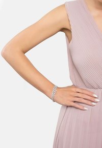 Silver bracelet with pear-shaped crystals, displayed on a slender wrist. The hand rests on a soft pink pleated dress. Smooth, elegant design.