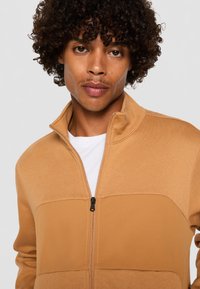 Brun zip-up jakke med tofarvet design, lavet af blødt stof med en struktureret overflade og en høj krave.