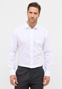 Camicia bianca a maniche lunghe con chiusura a bottoni, taschino sul petto, e bottoni a contrasto, abbinata a pantaloni scuri. Tessuto liscio.