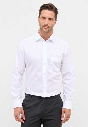 Witte langemouwen shirt met een knopenklep aan de voorkant, een enkele borstzak en contrasterende knopen, gecombineerd met donkere broek. Gladde stof.