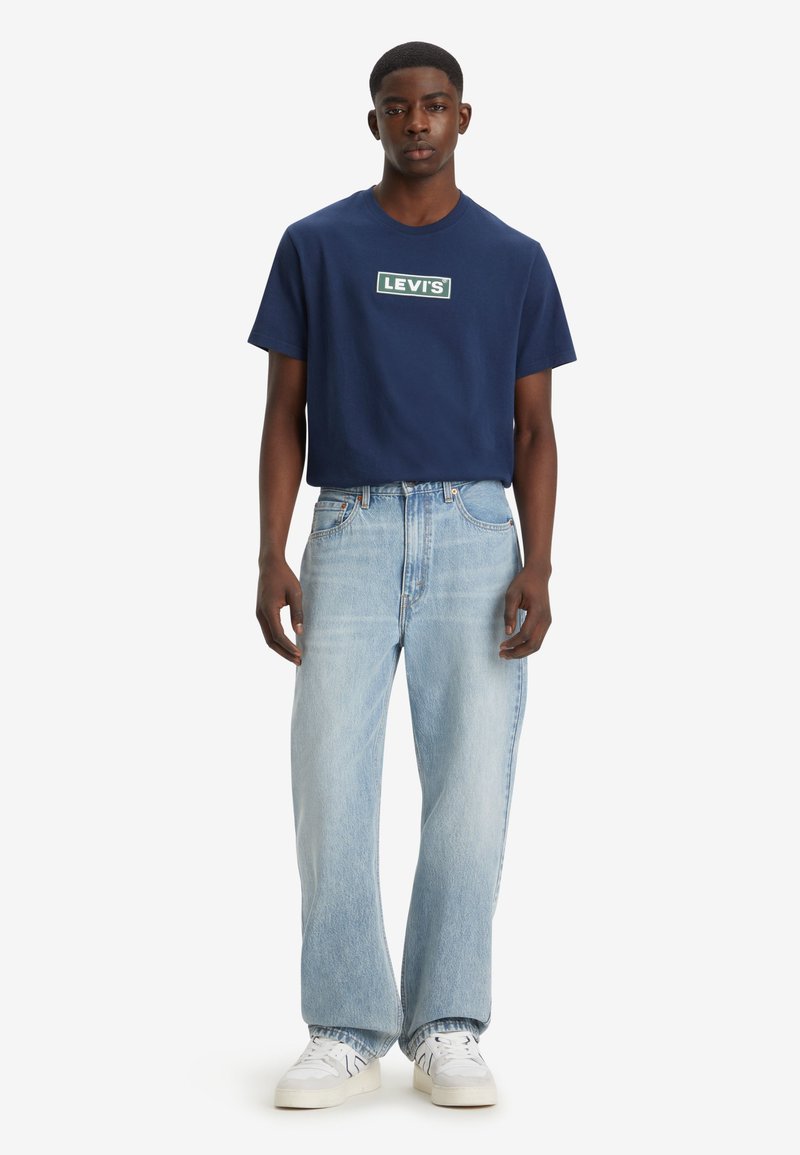 Man draagt een marineblauw Levi's T-shirt, lichtblauwe wijde spijkerbroek en witte sneakers, staand tegen een witte achtergrond.