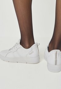 Zapatillas deportivas blancas de cuero texturizado, con diseño de cordones y perfil bajo. Presentan rayas azules y un acento plateado en el talón.