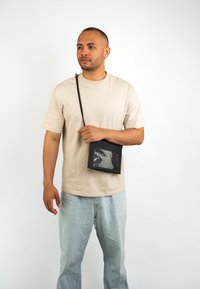 Fekete keresztpántos táska átlátszó elülső zsebbel, téglalap alakú, állítható pánttal, bézs pólóval és világoskék farmernadrággal viselve.