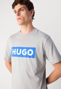 Pilkas medvilnės marškinėlis su mėlynu stačiakampiu grafiku ir baltu tekstu "HUGO." Trumpomis rankovėmis ir apvalia iškirpte. Lygus paviršius.