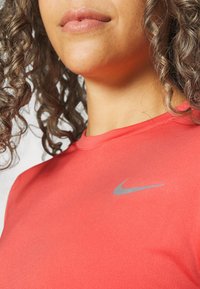 Kiharatukkainen nainen punainen Nike-urheilupaita yllään, näkyvissä ylävartalo ja alemman kasvojen osa, huulet neutraalit.