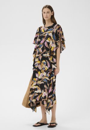 Žena v pestrofarebných šatách s abstraktným vzorom a volánovými rukávmi, drží veľkú béžovú tašku a má na nohách čierne žabky.