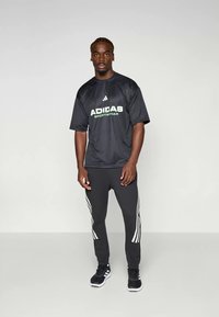 Zwart Adidas sportshirt met korte mouwen en groen logo, gecombineerd met zwarte broek met witte strepen aan de zijkanten en zwarte sportschoenen.