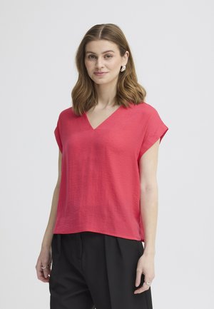 Donna con capelli castani fino alle spalle che indossa una camicetta rossa a maniche corte con scollo a V e pantaloni neri, in piedi davanti a uno sfondo chiaro uniforme.