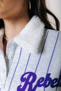 Maglione grigio con strisce verticali viola, colletto bianco peloso, ricamo "Rebel" in viola e chiusura a bottone al collo.
