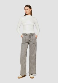 Weißes Langarmoberteil mit hohem Kragen, kombiniert mit grauen Denim-Hosen mit weitem Bein, die vordere Taschen und kontrastierende Nähte aufweisen. Bequeme Schuhe inklusive.