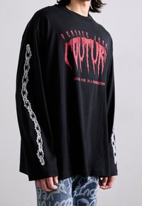 Musta pitkähihainen t-paita, jossa on punaiset "COUTURE"-tekstit ja valkeat ketjumaiset grafiikat hihoissa. Rentoa leikkausta, jossa pyöreä pääntie.