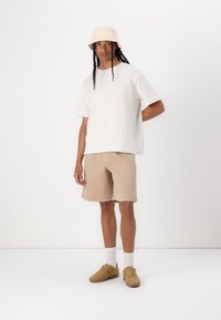 Witte katoenen t-shirt, beige shorts, beige emmerhoed en bruine sneakers. Comfortabele pasvorm, casual stijl, staand op een effen achtergrond.