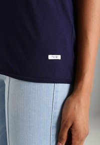 Detailní záběr osoby v námořnické modré košili s malou bílou nášivkou "Marc O'Polo Denim" a světle modrými džíny.