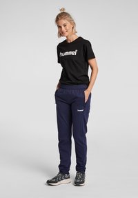 Svart T-shirt med "hummel"-logga, ihopparad med marinblå sweatpants som har sidofickor. Skorna är mörka med ljusa accenter.