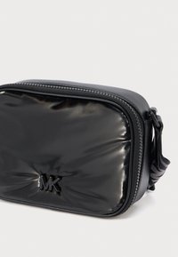 Μαύρη συνθετική τσάντα χιαστί με λείο, γυαλιστερό φινίρισμα, που διαθέτει φερμουάρ και λογότυπο στο μπροστινό μέρος. Περιλαμβάνει ρυθμιζόμενο λουρί.